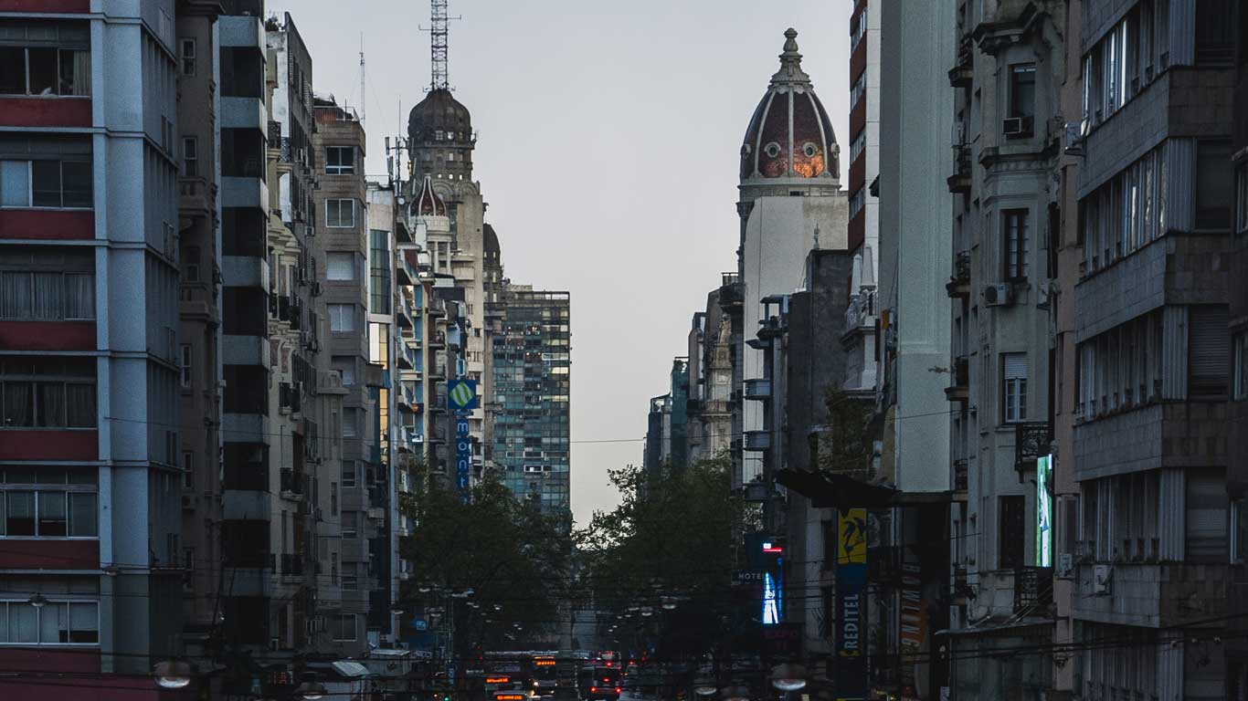 Vista da Avenida 18 de Julho em Montevidéu ao entardecer, cercada por edifícios altos e históricos. No horizonte, torres arquitetônicas se destacam, enquanto as luzes da cidade começam a brilhar. Esse é um excelente lugar onde ficar em Montevidéu.