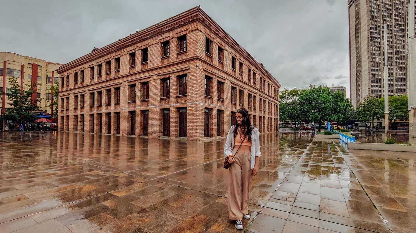 Mulher caminhando descontraída em uma praça da Candelaria em Medellín, em um dia de chuva. A mulher veste uma calça caqui, com top laranja e uma camisa cinza.