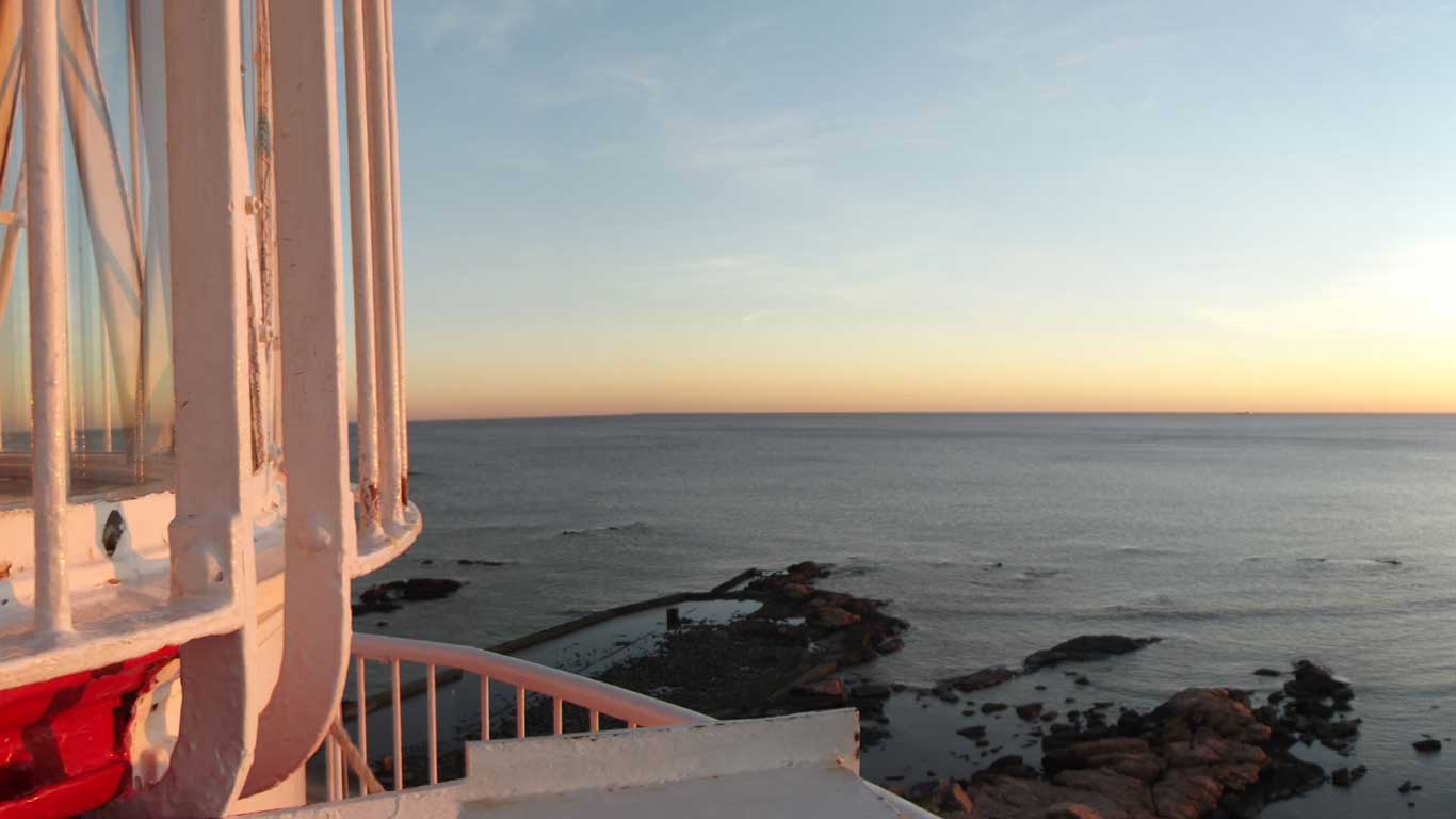 Imagem do Farol de Punta Carretas localizado à esquerda, capturada durante o pôr do sol. A estrutura branca e vermelha do farol está parcialmente visível, com o mar calmo ao fundo e algumas rochas espalhadas na beira da água. O céu está claro, com tons suaves de azul e laranja, característicos do fim de tarde, criando uma paisagem serena e encantadora.