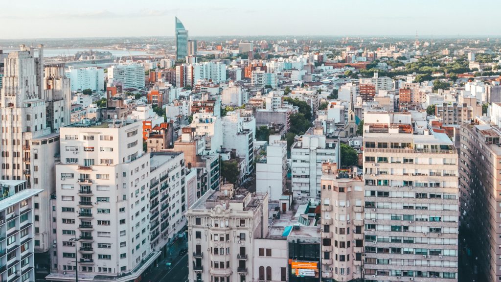 A imagem mostra uma vista aérea da cidade de Montevidéu, no Uruguai. A cena é composta por diversos prédios de diferentes tamanhos e estilos arquitetônicos, com a Rambla e o Rio da Prata visíveis ao fundo, destacando o horizonte urbano com uma mistura de edifícios antigos e modernos.