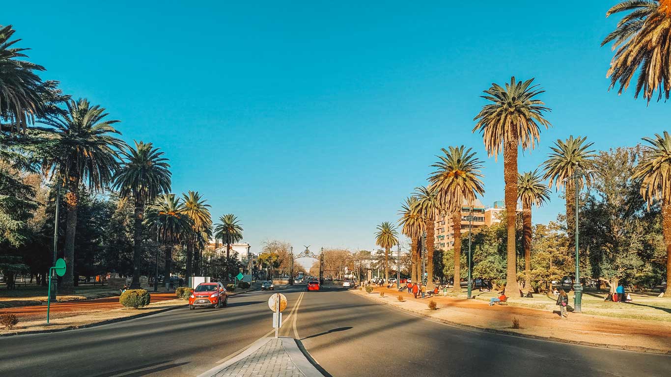 Avenida arborizada do Parque General San Martín, em Mendoza, Argentina, com palmeiras alinhadas dos dois lados da rua, pessoas caminhando e alguns carros ao longo da estrada, em um dia ensolarado.