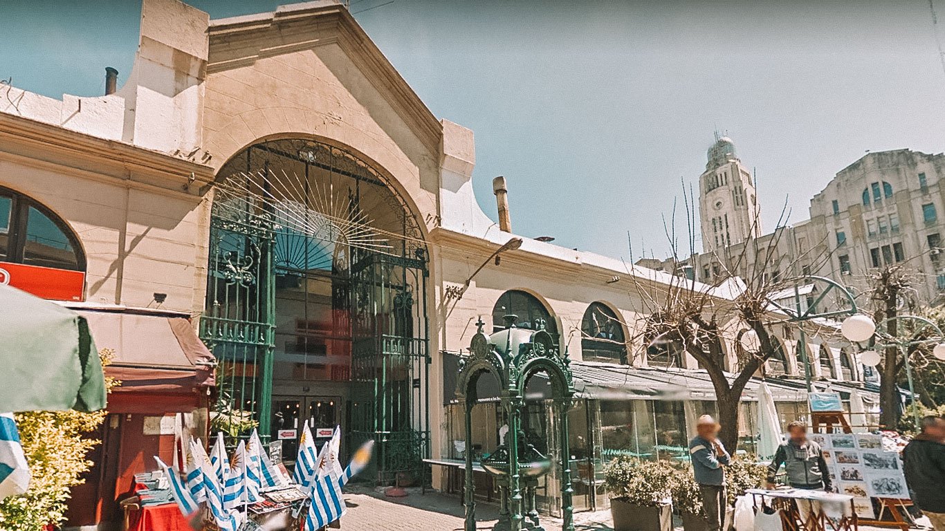 Entrada principal do Mercado del Puerto em Montevidéu, com sua arquitetura histórica destacada por um grande arco de ferro forjado. Na frente do mercado, há barracas com bandeiras do Uruguai e pessoas circulando.