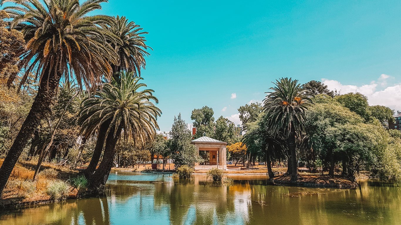 A imagem retrata uma parte do Parque Rodó, em Montevidéu, com um lago calmo cercado por palmeiras e vegetação exuberante. No fundo, é possível ver uma pequena estrutura retangular em estilo clássico, próxima à margem do lago. O céu está claro e azul, criando uma atmosfera relaxante em um dia ensolarado.