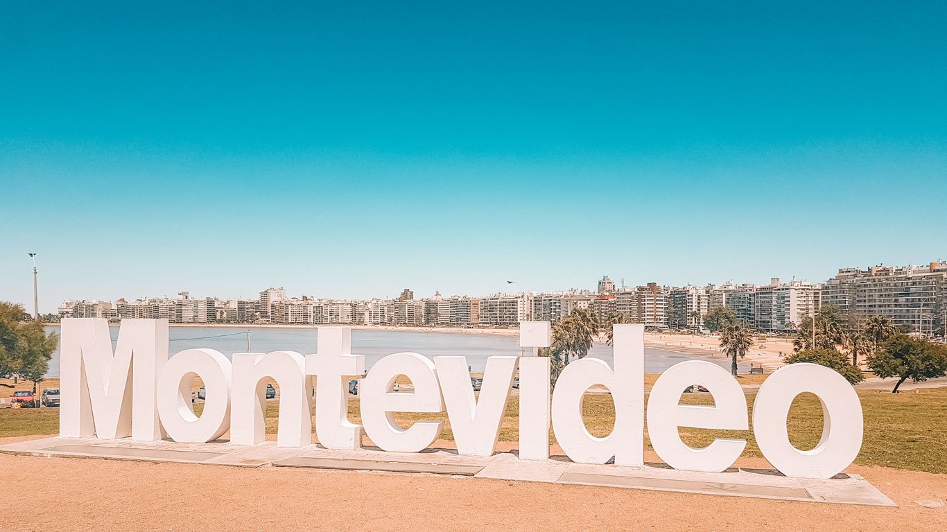 A imagem exibe o famoso letreiro "Montevideo" em grandes letras brancas, localizado na praia de Pocitos, com vista para a orla repleta de prédios residenciais ao fundo. O céu está claro e azul, e a praia aparece tranquila, destacando o charme costeiro da capital uruguaia.