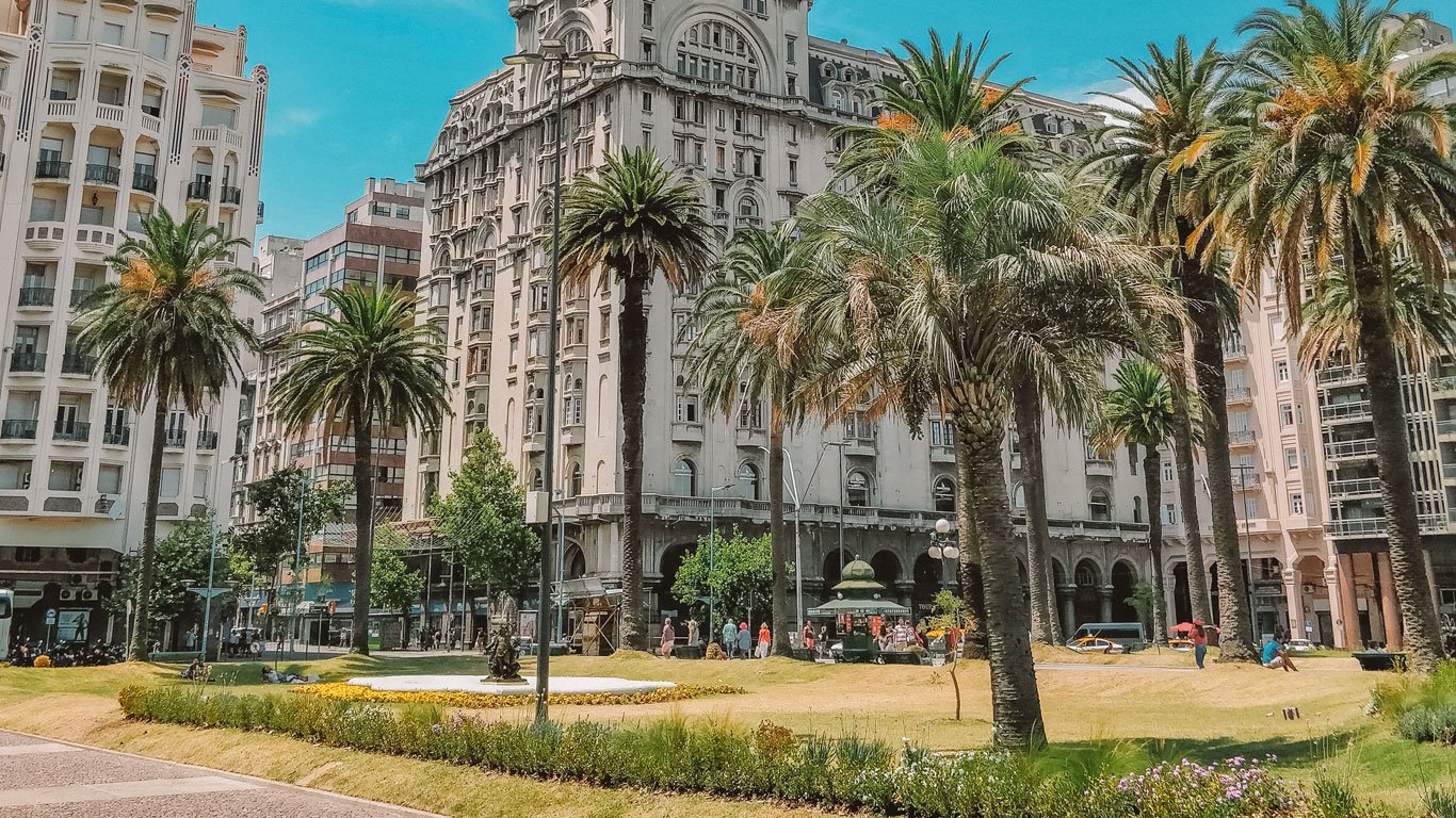 A imagem mostra a Praça Independência em Montevidéu, cercada por prédios históricos e modernos, como o icônico Palácio Salvo ao centro. Palmeiras altas decoram a praça, enquanto pedestres caminham e relaxam em um dia ensolarado. O cenário mistura arquitetura clássica e moderna, destacando-se como um dos principais pontos turísticos da cidade.