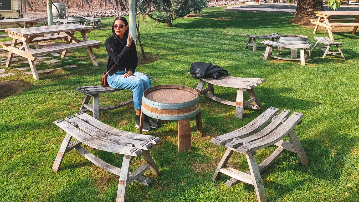 Mulher sentada em uma área verde na Bodega Tempus Alba em Mendoza, cercada por bancos de madeira rústicos e gramado verde, em um ambiente relaxante de vinícola.