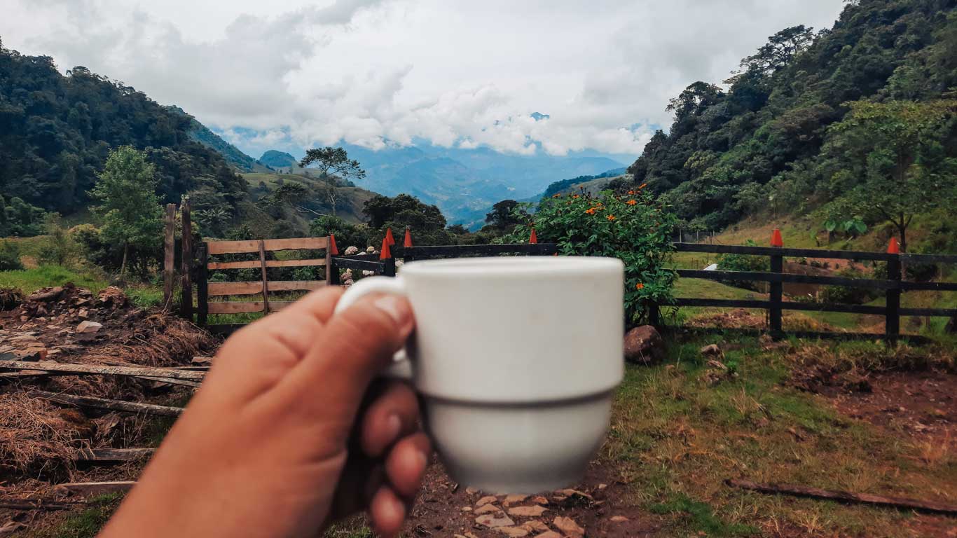 Mulher segurando uma xícara com água de panela, em frente a uma vista deslumbrante com morros cobertos de vegetação verde ao fundo, nos arredores de Jardín, Colômbia.