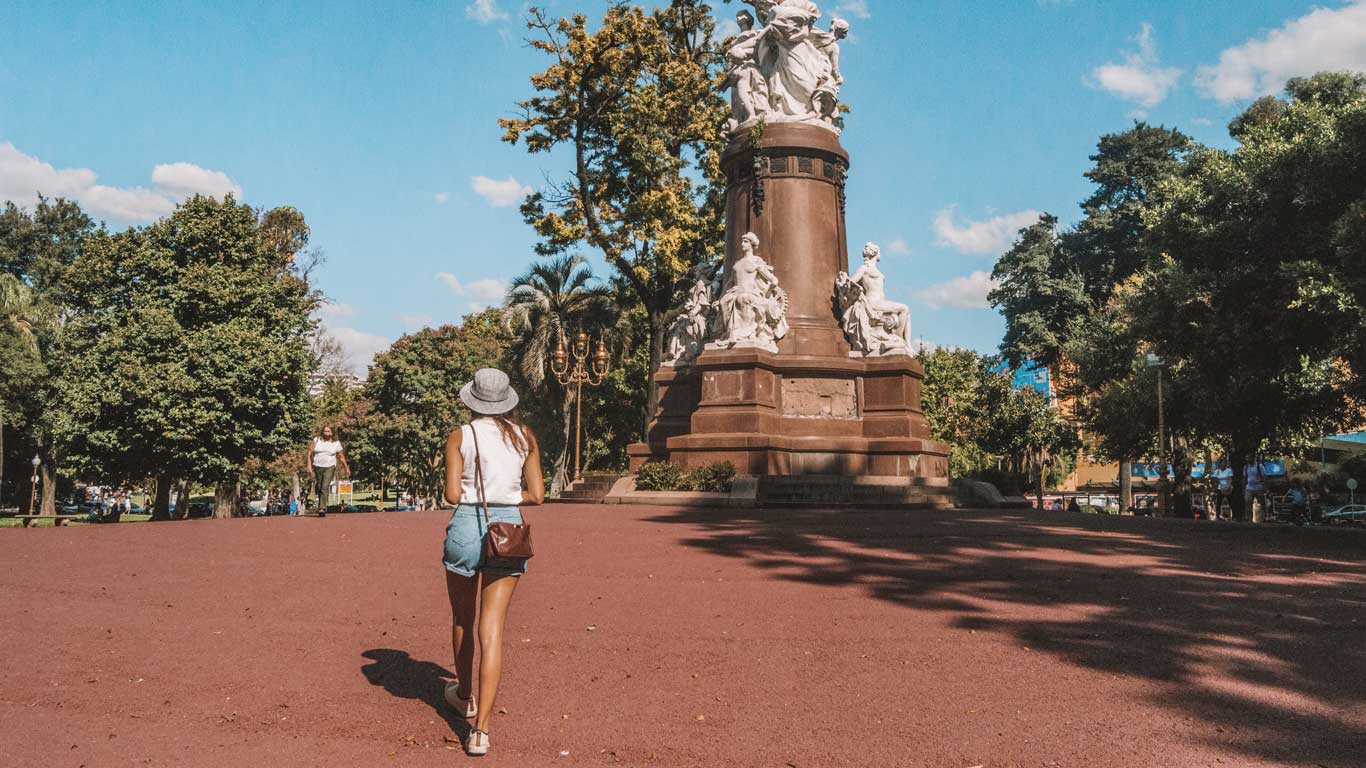 Mulher caminhando em direção a um grande monumento na Plaza Francia, em Buenos Aires. O monumento é adornado com estátuas detalhadas e está localizado em uma área arborizada, com céu azul e nuvens esparsas ao fundo. A mulher, vestida casualmente com chapéu e bolsa pequena, aprecia o ambiente tranquilo do parque enquanto caminha em direção à obra de arte.