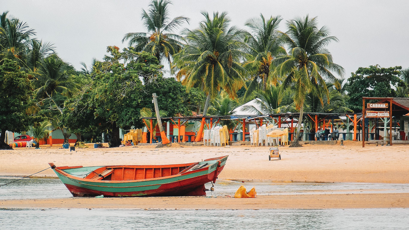 Praia de Coroa Vermelha, na Bahia, Brasil, com um barco de madeira colorido encalhado na areia próxima à água. Ao fundo, há barracas de praia com cadeiras empilhadas e mesas sob toldos, cercadas por palmeiras exuberantes.