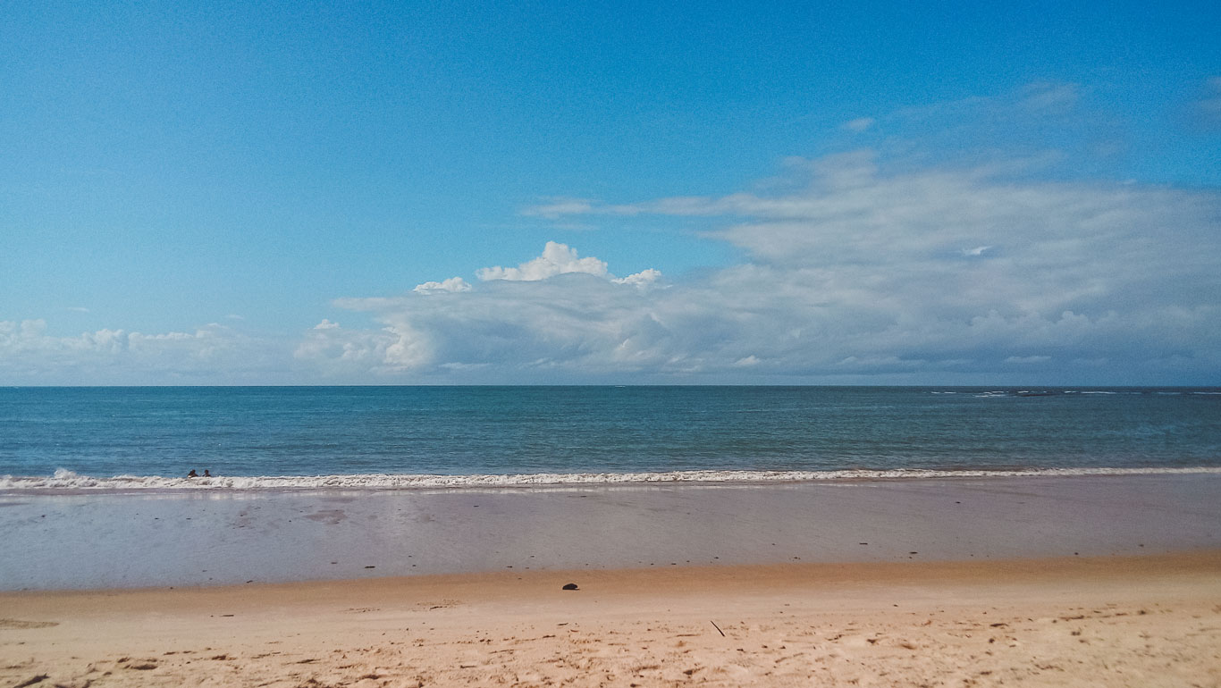 Praia de Curuípe, na Bahia, Brasil, com uma faixa de areia dourada e o mar tranquilo em tons de azul. Duas pessoas podem ser vistas nadando nas águas calmas, enquanto o céu azul com algumas nuvens complementa o cenário sereno.