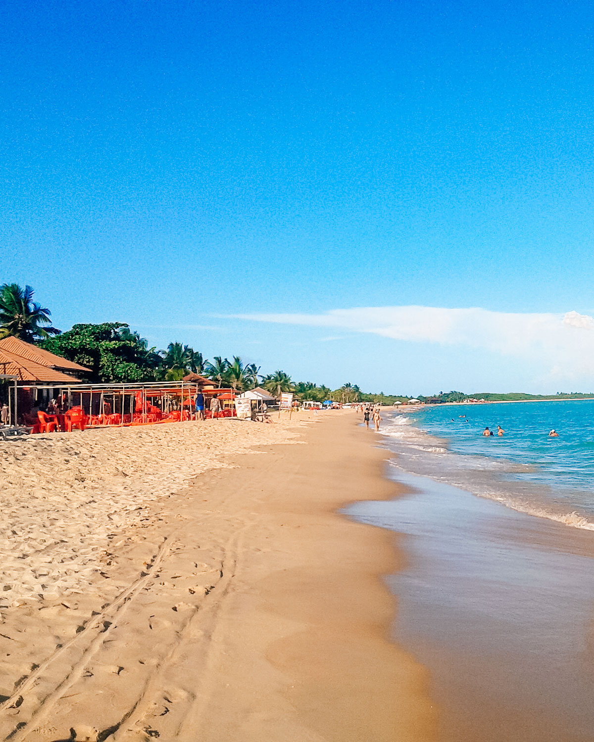 Vista da Praia de Taperapuã, a melhor praia onde ficar em Porto Seguro, Bahia. A praia tem areia dourada, algumas pessoas nadando no mar e uma fileira de barracas ou restaurantes à beira-mar, com cadeiras e guarda-sóis vermelhos. Ao fundo, há palmeiras e um céu azul claro, criando uma atmosfera tropical e relaxante.