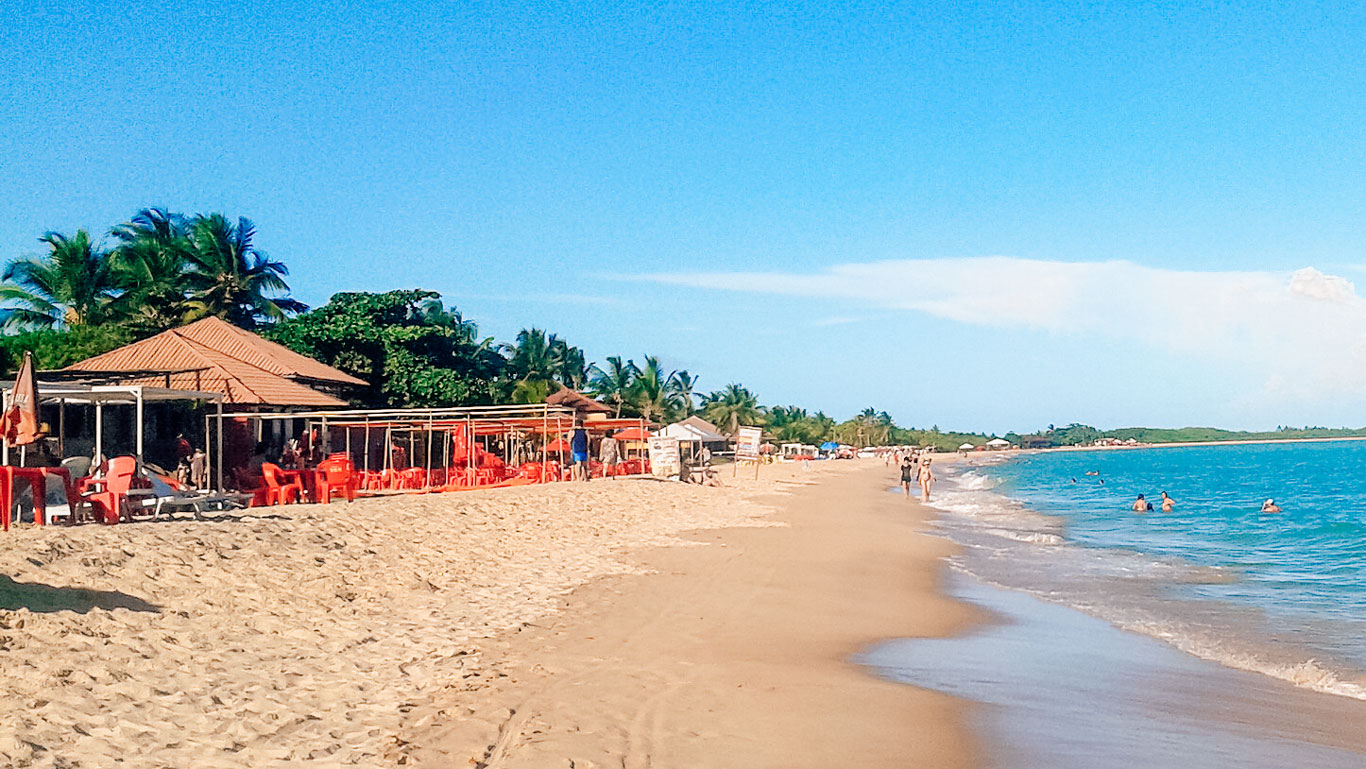 Vista da Praia de Taperapuã, a melhor praia onde ficar em Porto Seguro, Bahia. A praia tem areia dourada, algumas pessoas nadando no mar e uma fileira de barracas ou restaurantes à beira-mar, com cadeiras e guarda-sóis vermelhos. Ao fundo, há palmeiras e um céu azul claro, criando uma atmosfera tropical e relaxante.