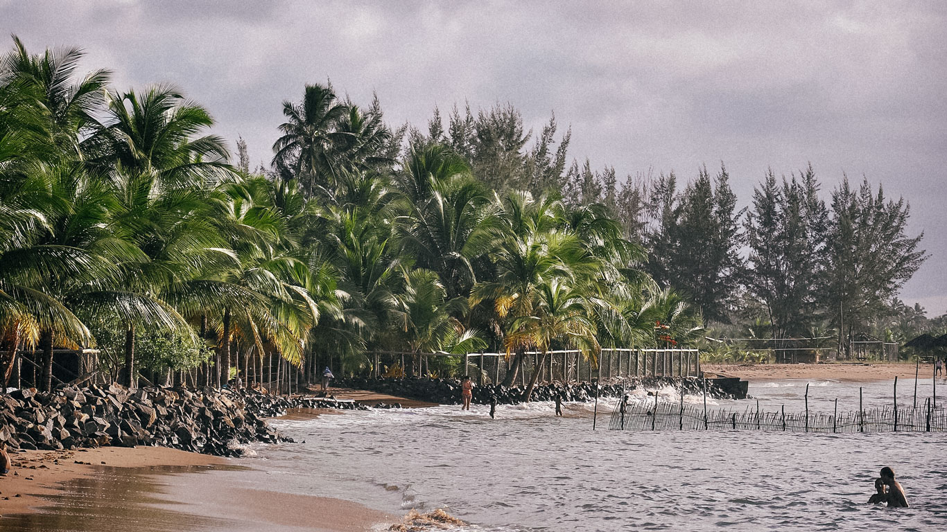 Imagem da Praia do Mutá, na Bahia. Ela destaca uma orla cheia de vegetação com algumas pessoas no mar.