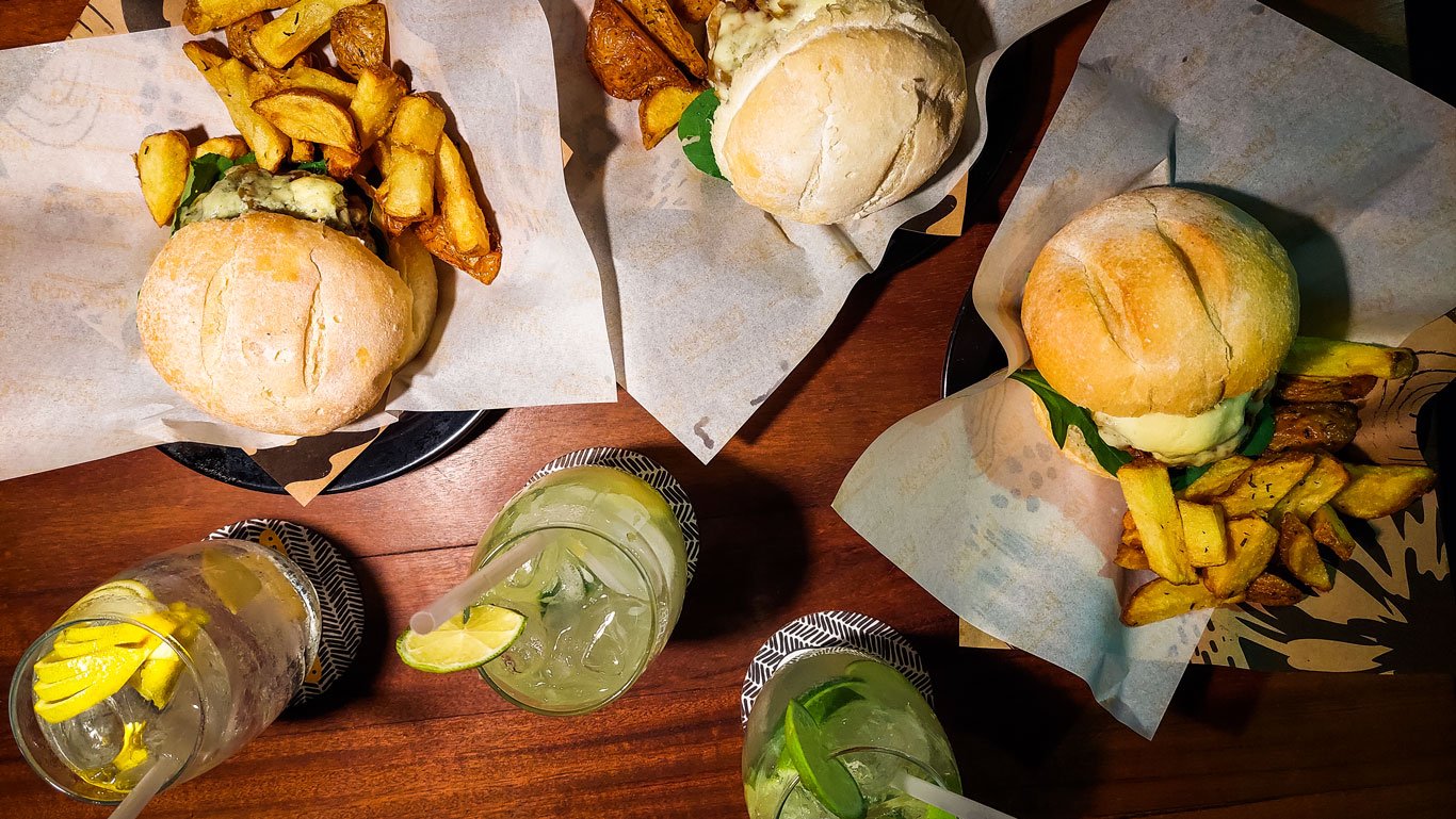 Visão aérea da mesa do Van Gogh Hamburgueria, com três hambúrgueres servidos em pães redondos, acompanhados de batatas fritas rústicas. Ao lado dos pratos, há copos de caipirinhas decorados com rodelas de limão e canudos.