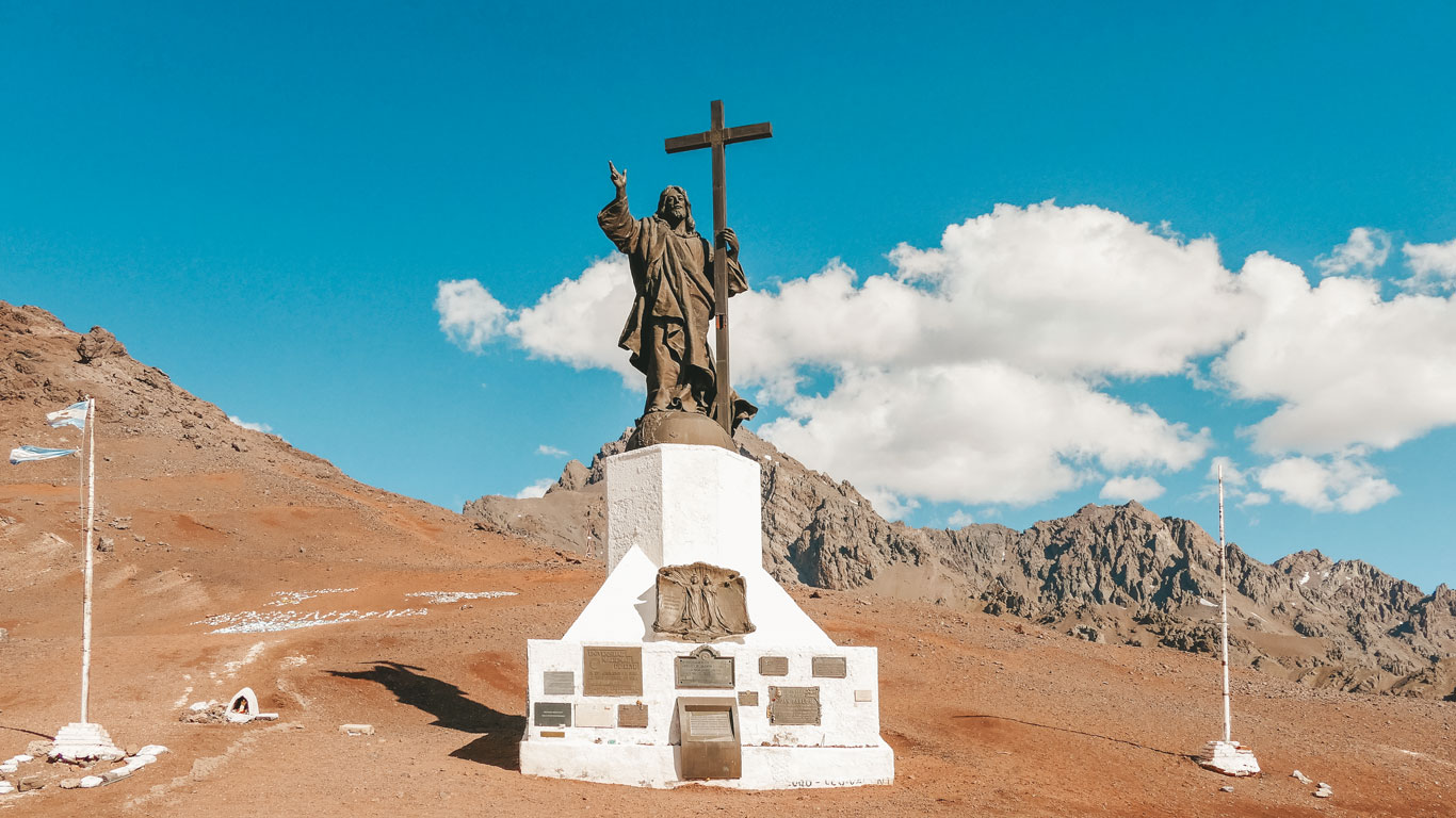 Imagem do monumento Cristo Redentor dos Andes, localizado na fronteira entre a Argentina e o Chile, em uma região montanhosa. A estátua de bronze de Jesus Cristo segura uma cruz com uma mão enquanto faz um gesto de bênção com a outra. A base branca do monumento contém várias placas comemorativas. À esquerda e à direita, há mastros com bandeiras argentinas, balançando suavemente ao vento.