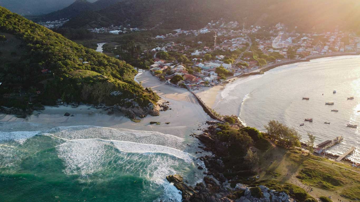 Vista aérea da baía da Praia da Armação, uma praia tranquila onde ficar em Florianópolis, cercada por montanhas verdes, com uma pequena vila e um cais ao lado da praia, onde o mar encontra a areia clara.