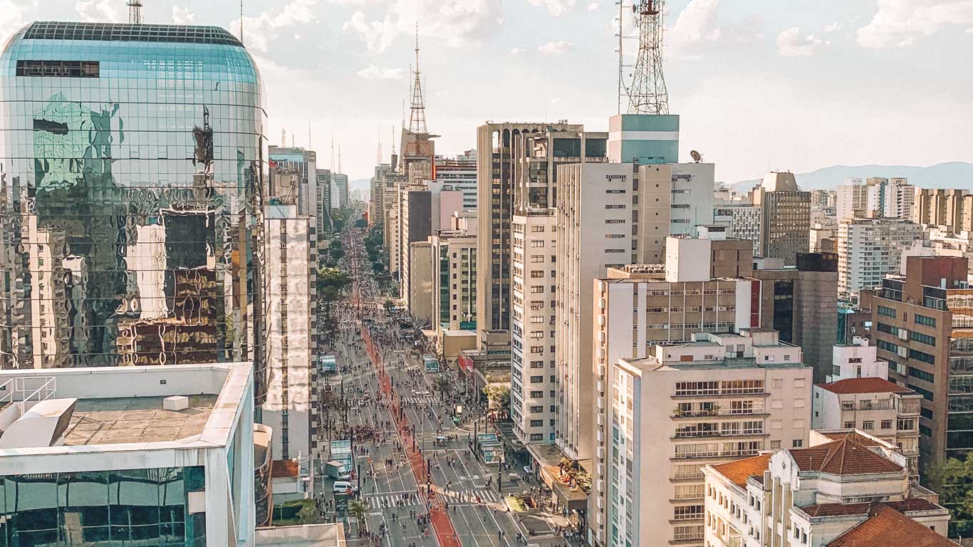 Vista aérea panorâmica da Avenida Paulista em São Paulo, Brasil, com arranha-céus alinhados na rua e uma atmosfera urbana movimentada sob um céu parcialmente nublado.