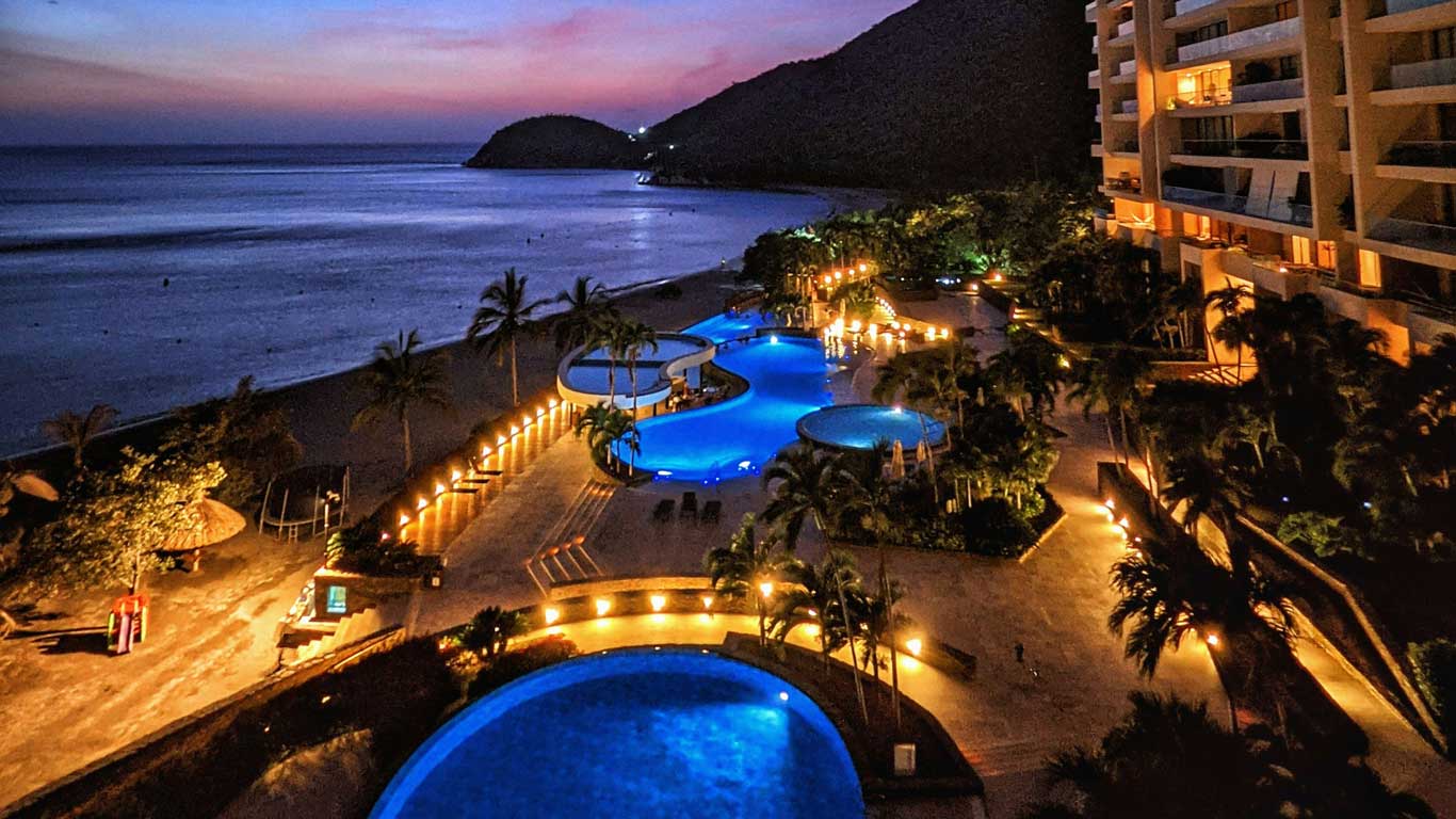 Vista noturna de um resort em frente à praia em Bello Horizonte, Santa Marta, com várias piscinas iluminadas e palmeiras ao redor. Ao fundo, é possível ver a praia e montanhas sob um céu colorido pelo pôr do sol.