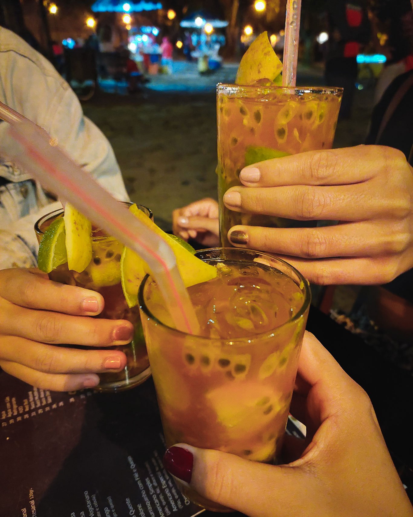 Três mãos levantando drinks Jorge Amado, em um ambiente noturno na Cachaçaria Cana da Praça. O fundo está suavemente desfocado, com luzes brilhantes.