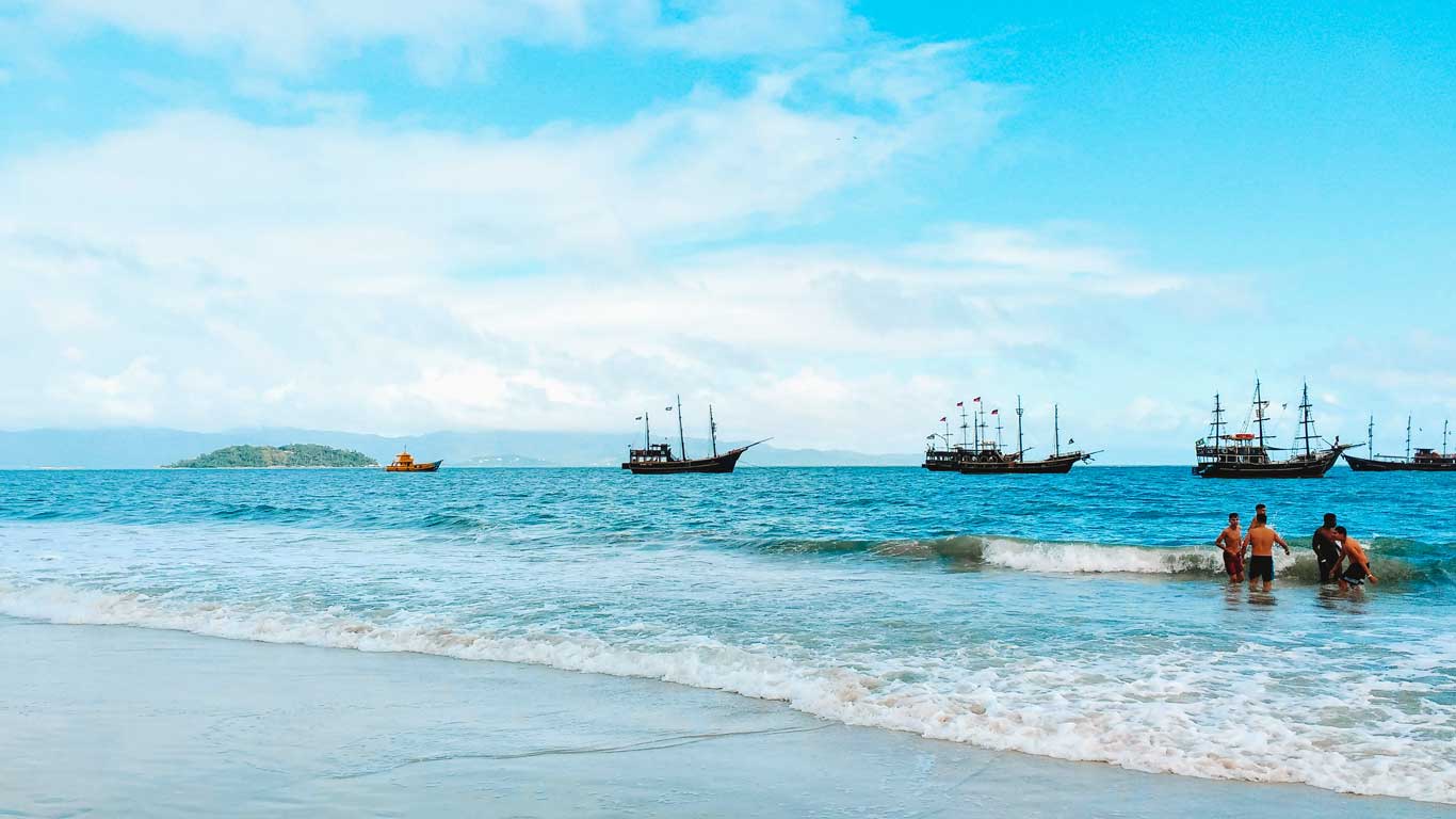 Praia de Canasvieiras, uma bos opção de onde ficar em Florianópolis com a família, com águas calmas, onde pessoas estão na água próximo à costa e, ao fundo, há várias embarcações de estilo antigo, como galeões, ancoradas.