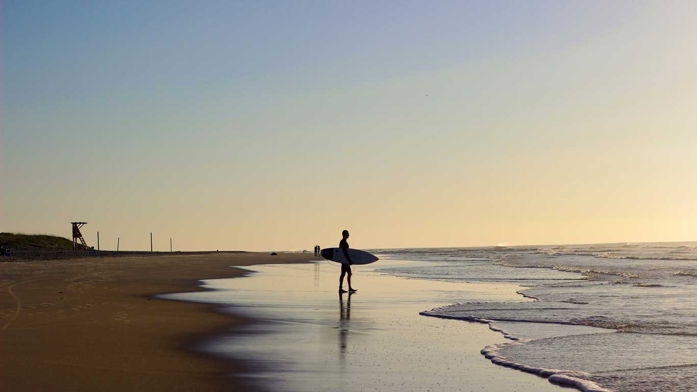 Um surfista em pé na beira da praia do Campeche em Florianópolis, ao pôr do sol, segurando sua prancha de surfe, enquanto o mar toca suavemente a areia ao fundo.