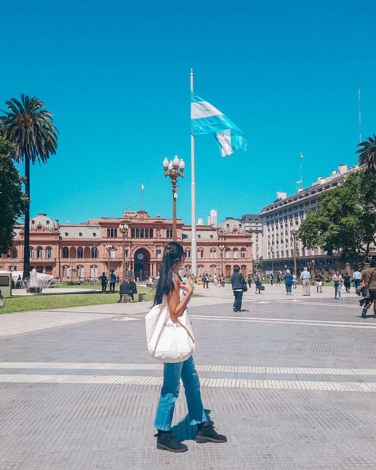 A imagem mostra a Plaza de Mayo com a Casa Rosada ao fundo, sob um céu azul claro. Uma grande bandeira da Argentina está hasteada ao centro, e várias pessoas caminham pela praça. Em primeiro plano, uma mulher com uma bolsa grande caminha despreocupadamente pela área pavimentada.