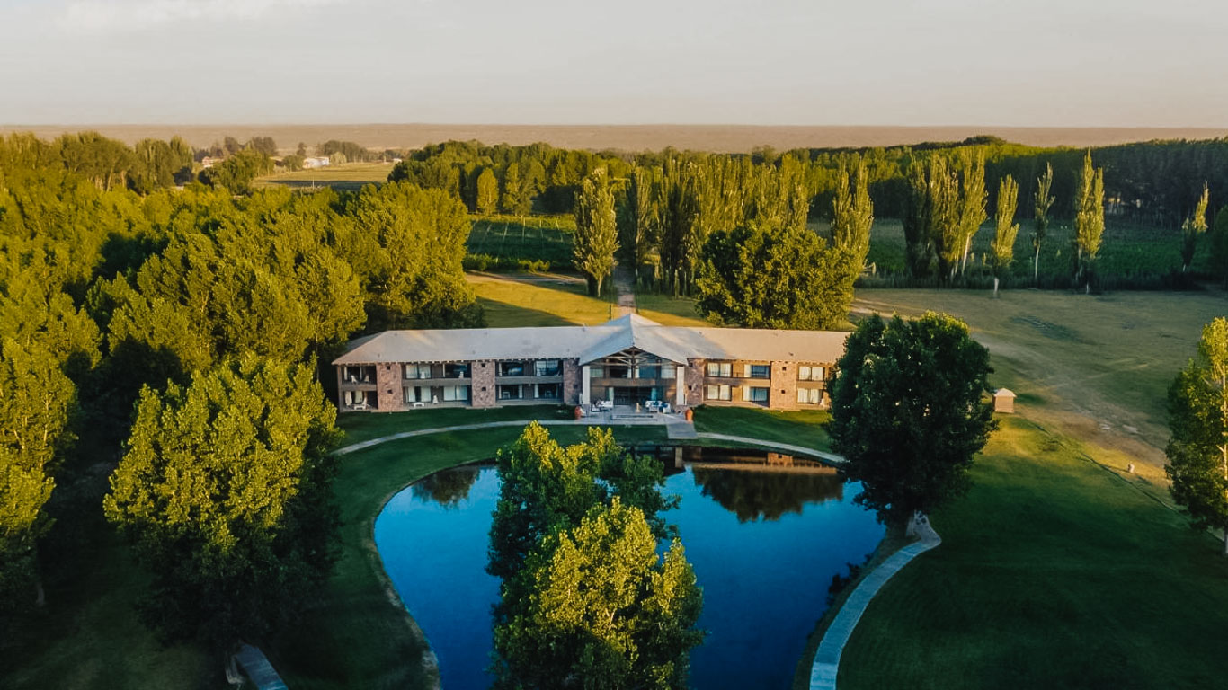 Imagem aérea de uma propriedade cercada por uma densa área verde em Mendoza, Argentina. No centro, há um grande edifício de estilo rústico, com dois andares e uma fachada de tijolos, localizado em frente a um pequeno lago que reflete a paisagem ao redor. Árvores altas e exuberantes cercam a construção, criando um ambiente tranquilo e isolado.