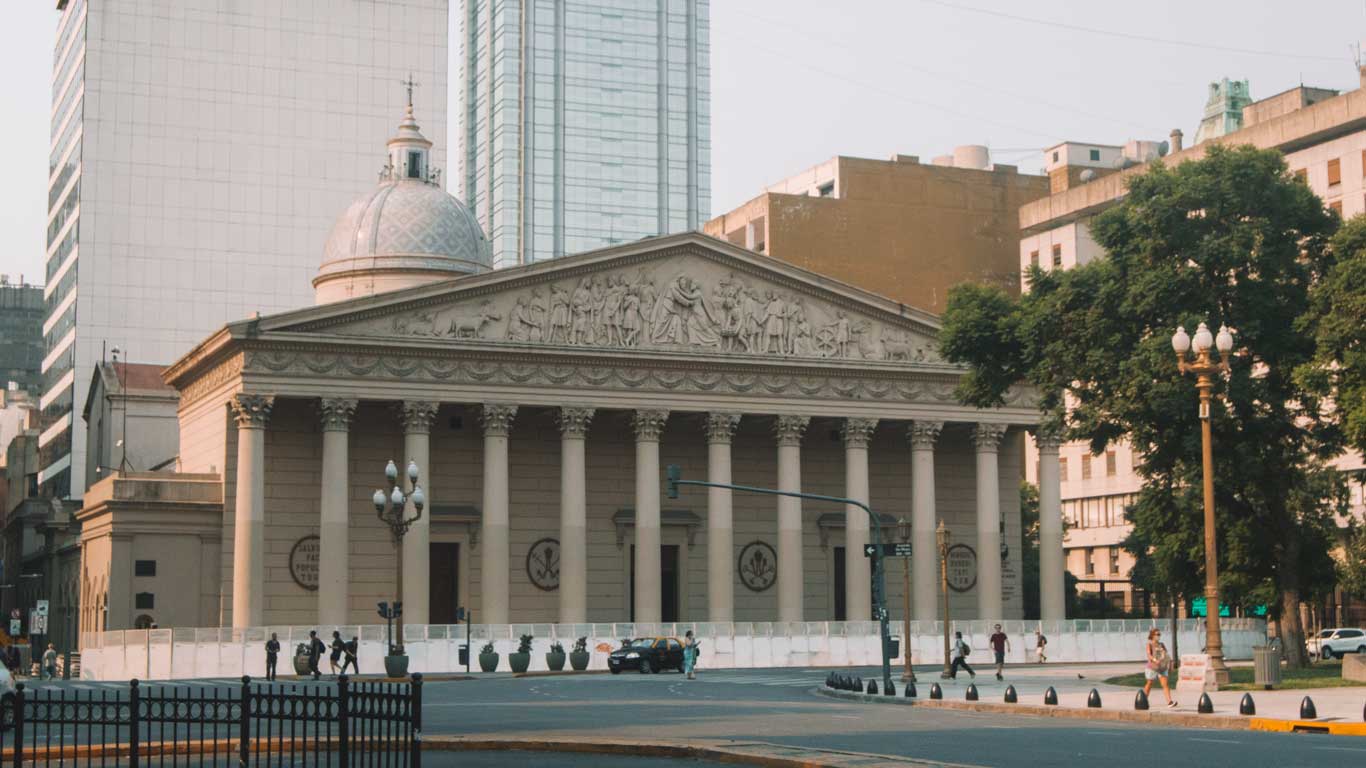 A imagem mostra a fachada da Catedral Metropolitana de Buenos Aires, com sua arquitetura neoclássica destacada por colunas altas e um frontão adornado com relevos detalhados. Ao fundo, há prédios modernos que contrastam com o estilo clássico da catedral. Algumas pessoas caminham pela calçada em frente, enquanto um carro passa pela rua.