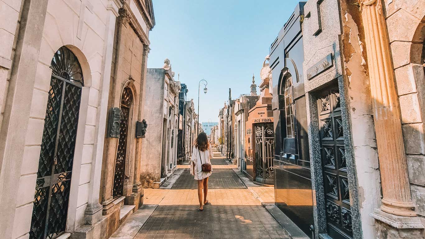 Mulher caminhando por entre túmulos no Cemitério da Recoleta, em Buenos Aires. As sepulturas, com suas fachadas elaboradas em mármore e pedra, alinham-se ao longo do estreito caminho.