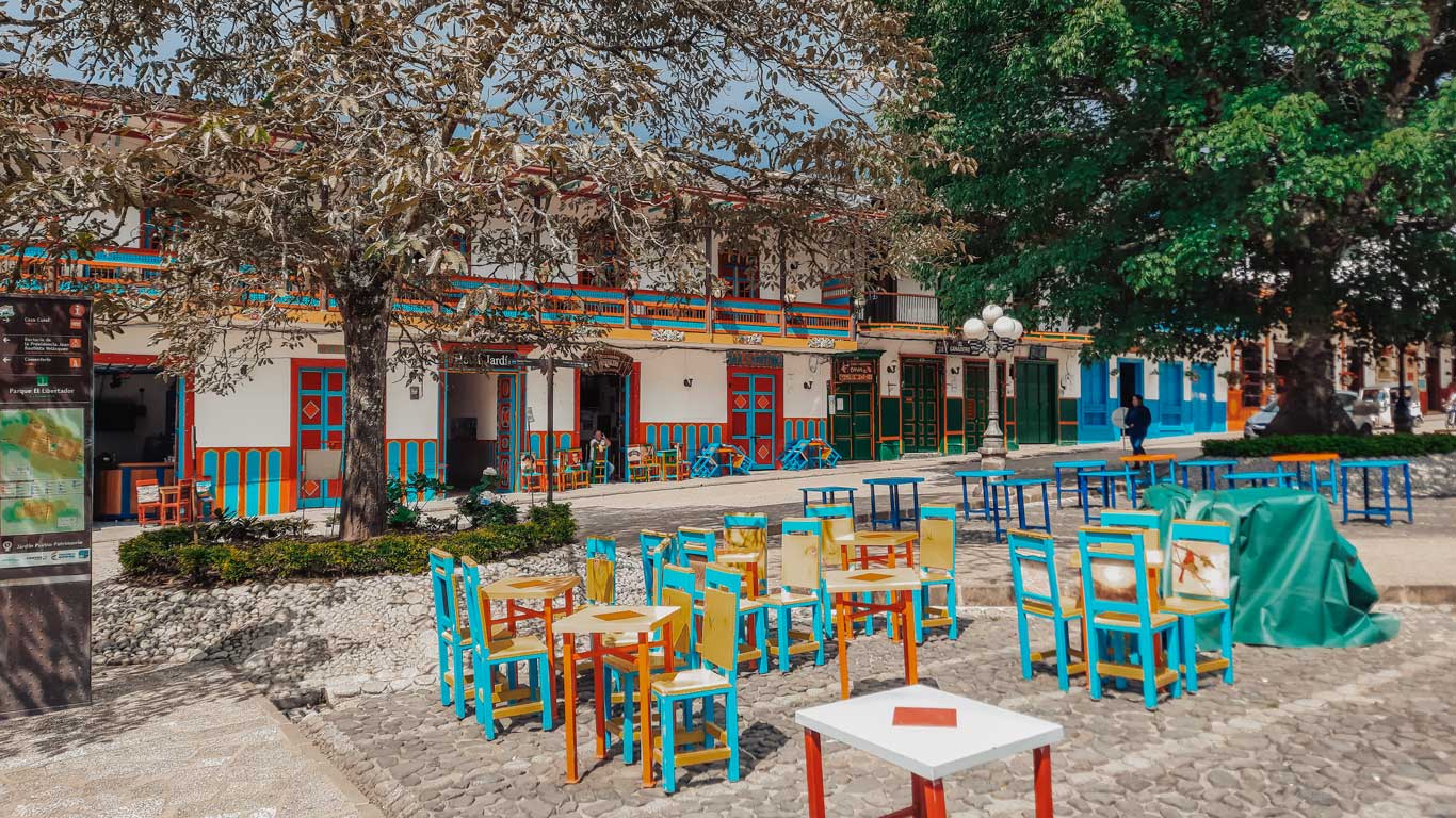 Mesas e cadeiras coloridas em tons de amarelo, vermelho e azul, dispostas na praça e na calçada dos bares e restaurantes que ficam no entorno da praça, o melhor lugar onde ficar em Jardín.