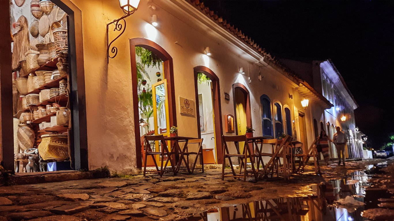 Charmosa rua de paralelepípedos no centro de Paraty à noite, com edifícios coloniais brancos iluminados por postes de luz. À esquerda, uma loja exibe uma coleção de cestos artesanais tradicionais.