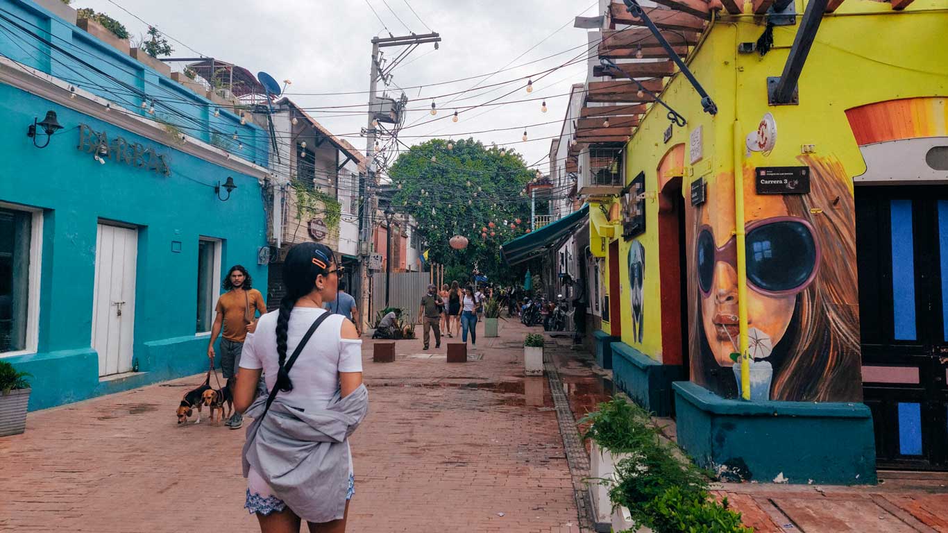 Rua de pedestres no centro histórico de Santa Marta, com edifícios coloridos de um lado azul e do outro amarelo com murais vibrantes. Pessoas caminham pela rua sob uma decoração de luzes penduradas, e há uma sensação de atmosfera descontraída e artística no local.