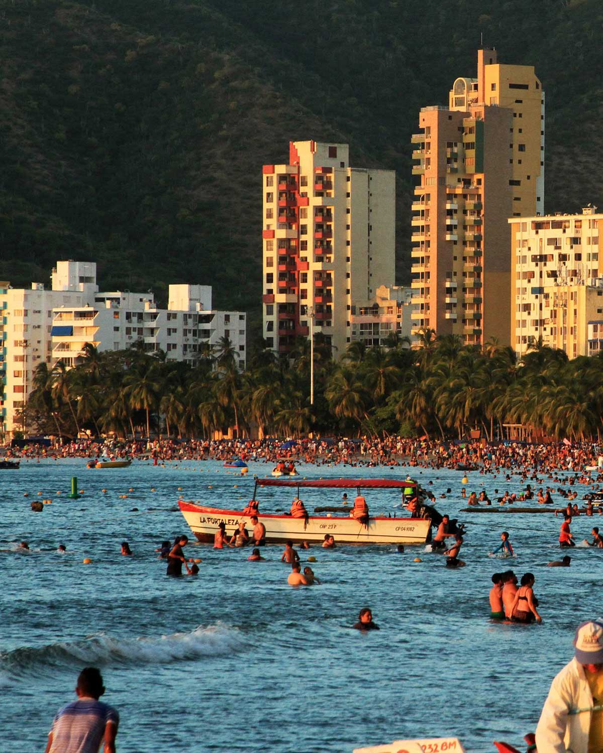 Praia de El Rodadero em Santa Marta, cheia de pessoas aproveitando o mar. Ao fundo, há prédios altos com montanhas verdejantes atrás, formando um contraste com as palmeiras na orla.