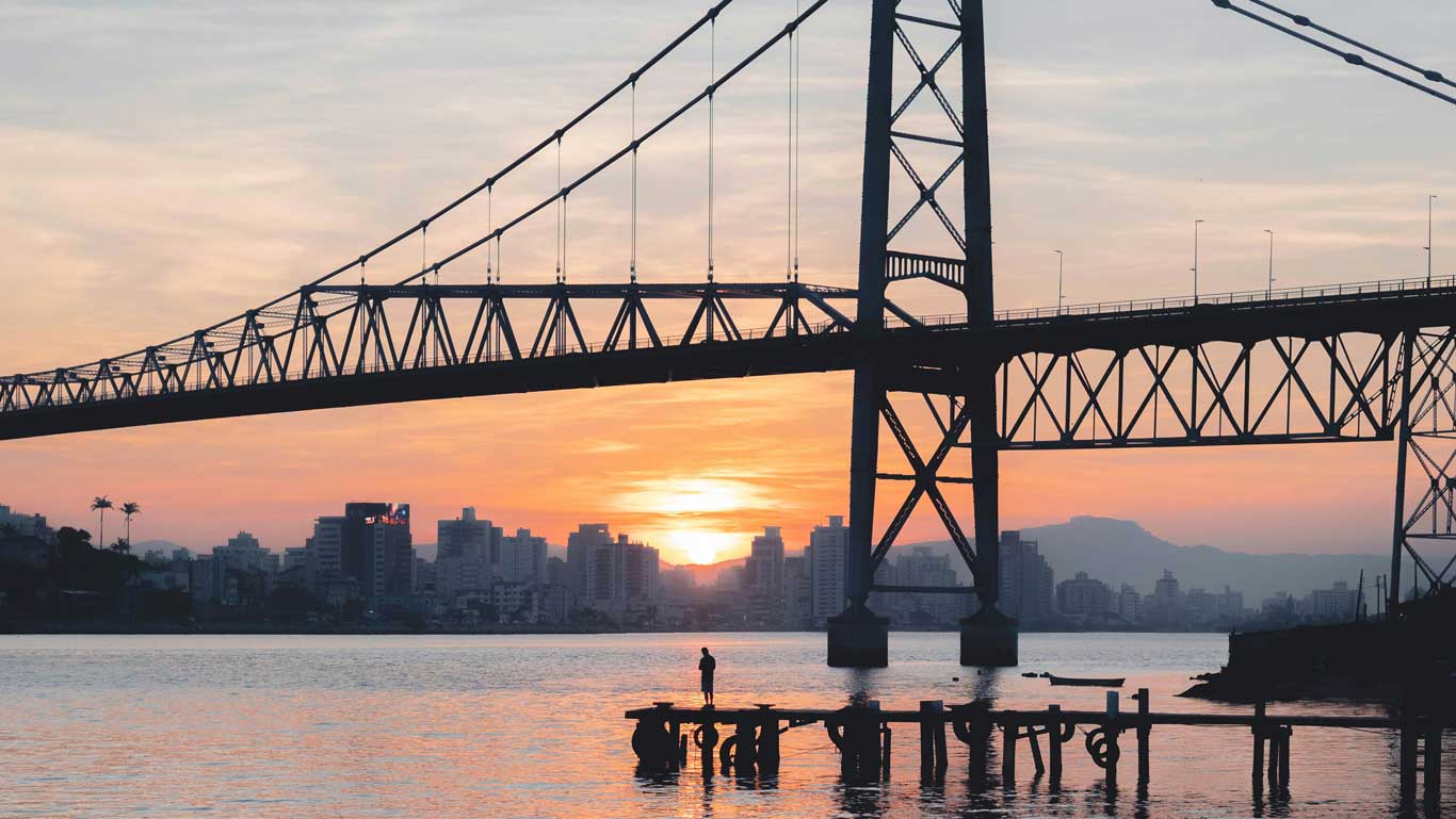 Ponte Hercílio Luz ao entardecer, ligando a ilha à cidade de Florianópolis, com um lindo pôr do sol e prédios no horizonte.