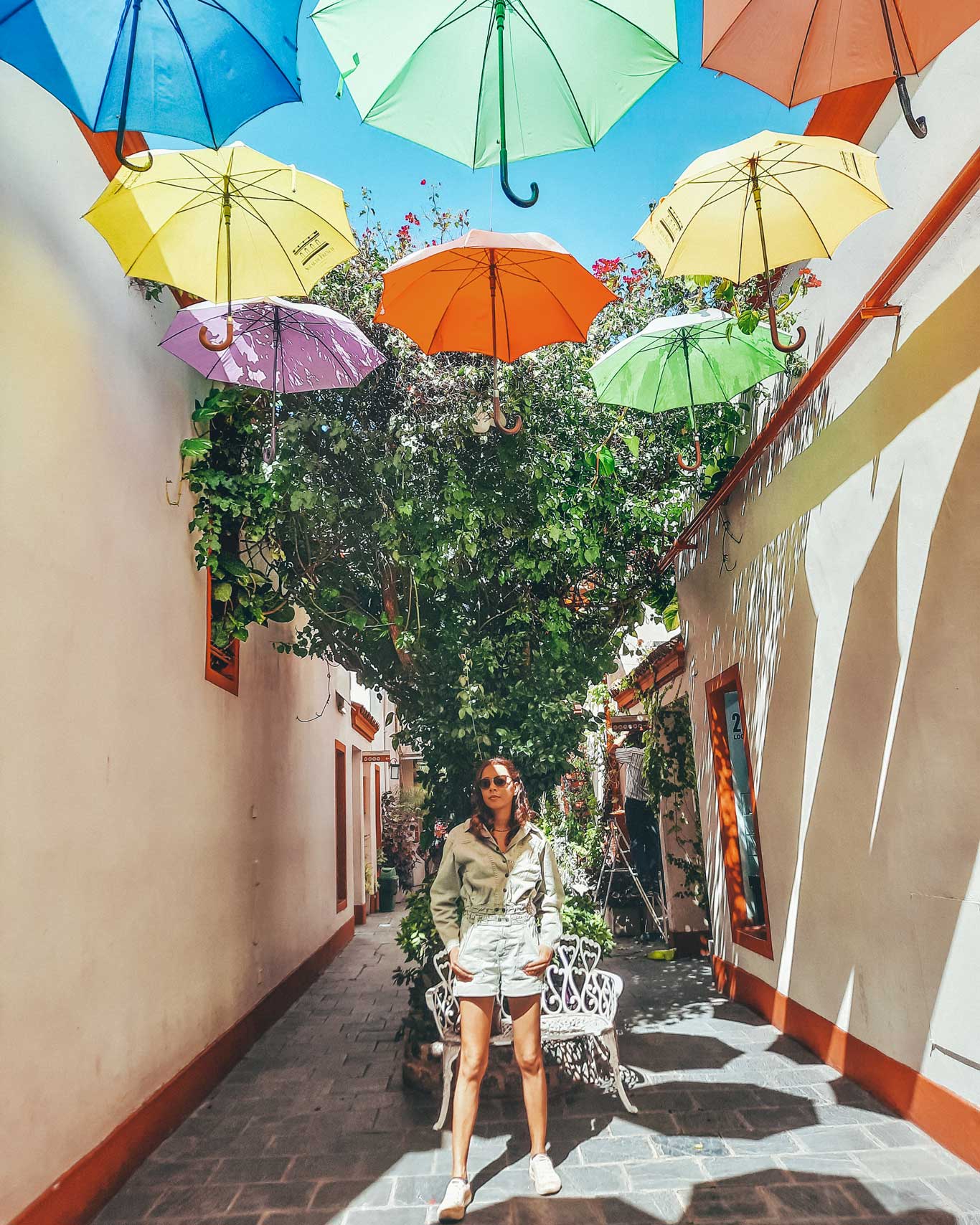 Mulher em uma passagem charmosa na Galeria Salar French, em Buenos Aires, onde guarda-chuvas coloridos estão suspensos acima, criando uma decoração vibrante. O ambiente é cercado por plantas e bancos decorativos de ferro, com paredes claras e detalhes em tons de laranja.