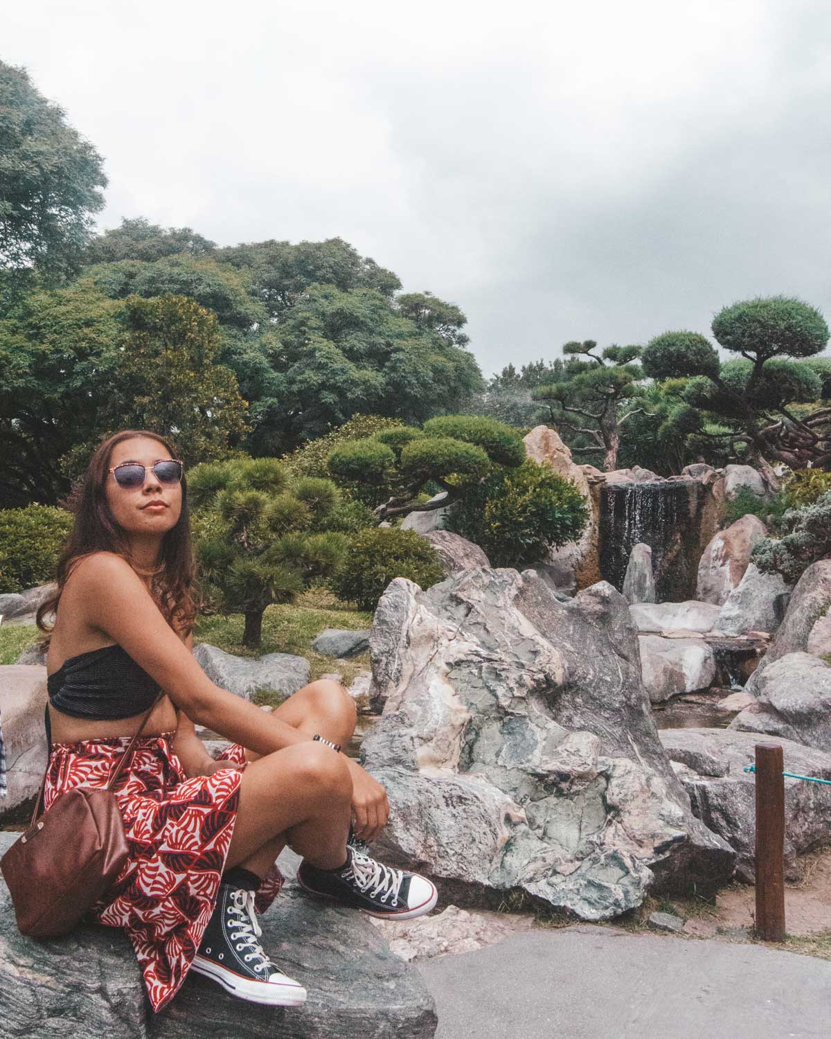 Mulher sentada em uma pedra no Jardim Japonês de Buenos Aires, cercada por árvores podadas e rochas típicas de jardins orientais. Ao fundo, há uma pequena cachoeira fluindo entre as pedras, criando um cenário sereno e natural. A mulher, usando óculos de sol e um estilo casual.