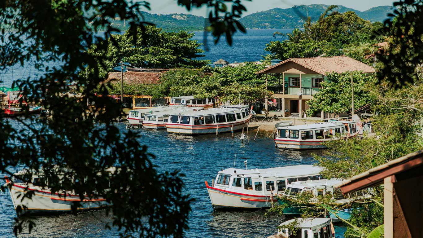 Pequenas embarcações ancoradas em uma marina na Lagoa da Conceição, rodeada de vegetação e casas simples, com o mar azul ao fundo e montanhas à distância.