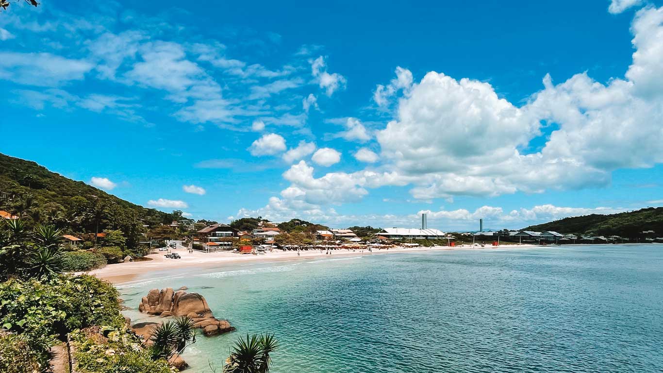 Vista da Praia da Lagoinha, uma tranquila com águas cristalinas e casas ao longe, margeada por vegetação e montanhas sob um céu azul.