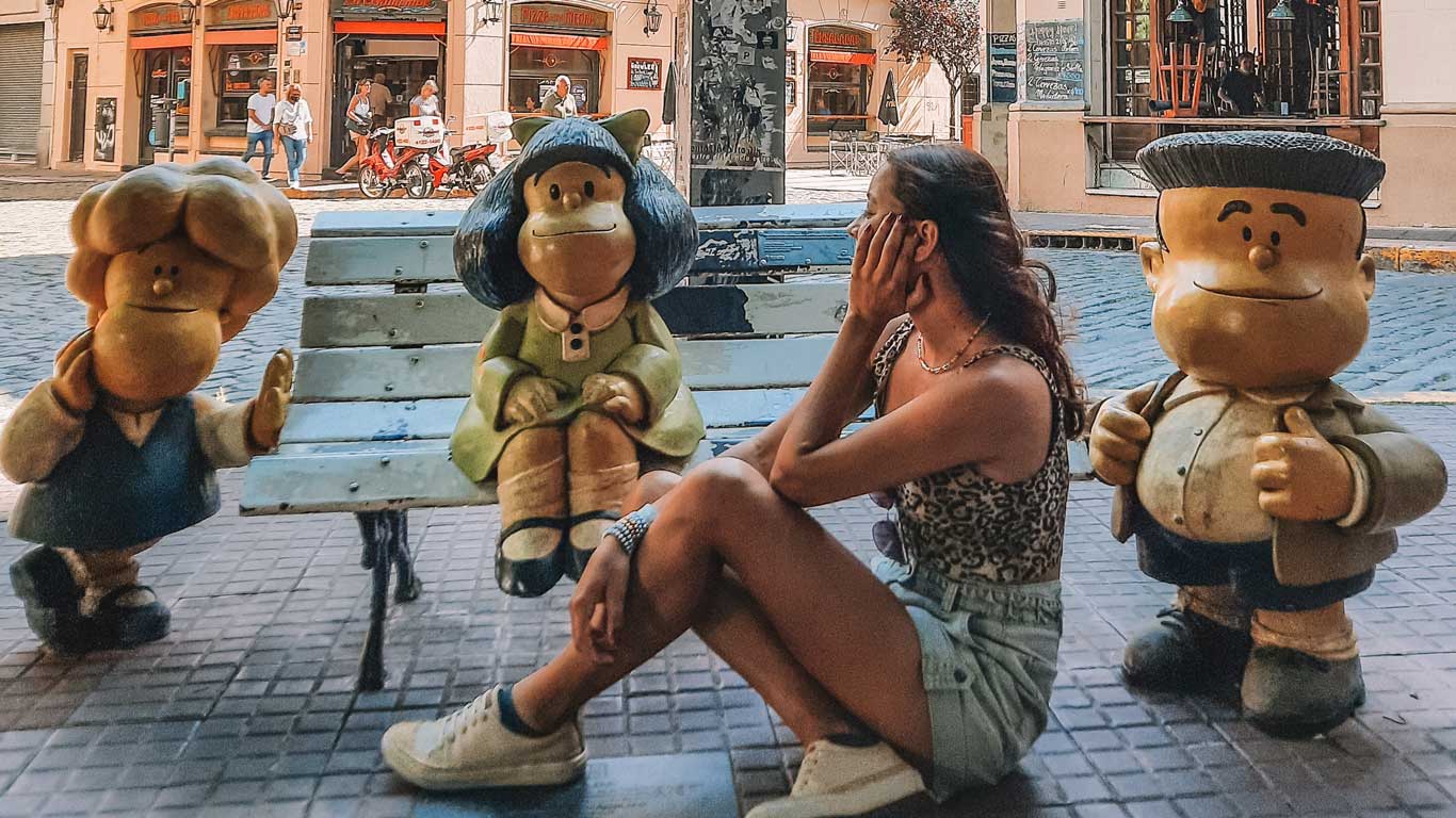 Mulher sentada ao lado das famosas estátuas dos personagens da Mafalda, localizadas em San Telmo, Buenos Aires. A personagem principal, Mafalda, está sentada em um banco com seus amigos Manolito e Susanita ao seu lado, enquanto a mulher parece interagir de forma divertida com as esculturas.