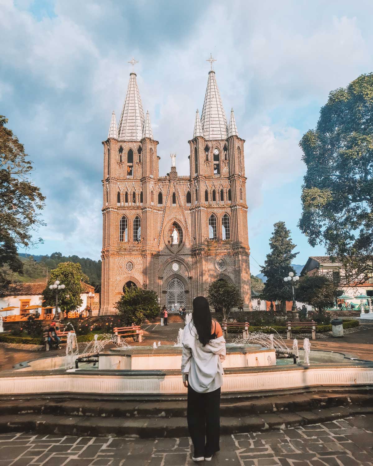 Mulher de cabelos escuros, camisa branca e calça preta, posando para uma foto de costas em frente a Catedral de Jardín, Colômbia. Ao fundo, a Catedral grandiosa com suas torres, localizada em frente a uma praça no centro do pueblo, o melhor lugar para se hospedar em Jardín.