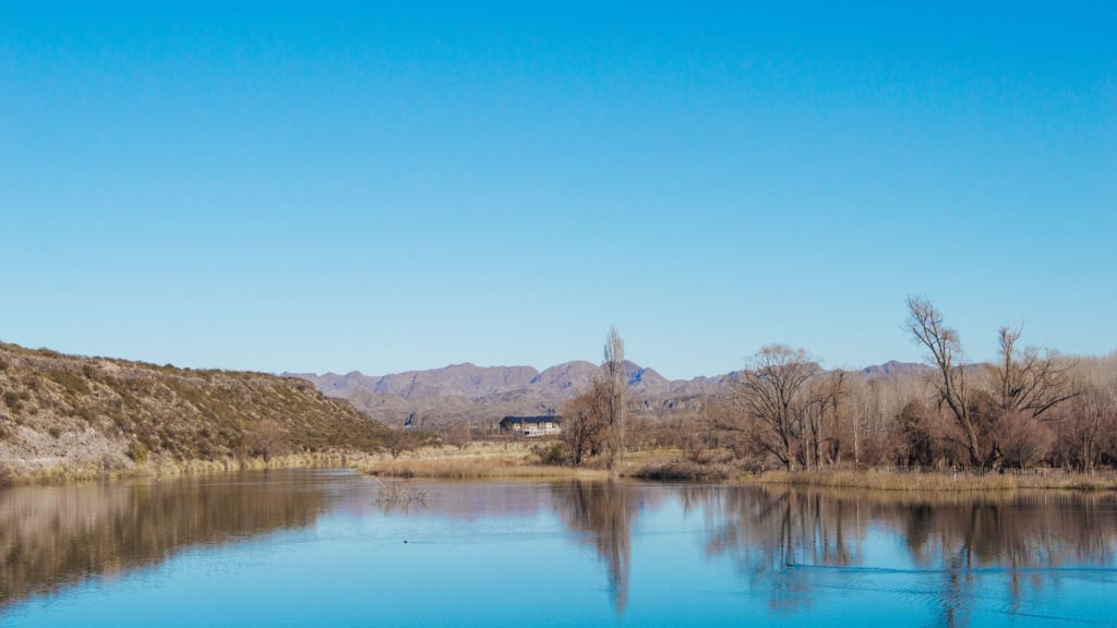 Foto de uma paisagem natural em Mendoza, Argentina, com um lago refletindo o céu azul claro e árvores secas ao redor. Ao fundo, é possível ver uma cadeia de montanhas de cor acinzentada e uma construção no meio da paisagem, possivelmente um edifício rústico ou pousada. O cenário transmite tranquilidade, com vegetação árida e sem folhas típicas do outono.