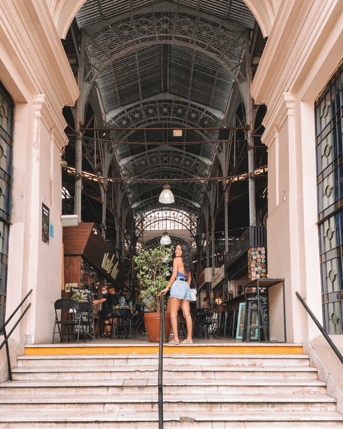 Mercado de San Telmo, em Buenos Aires, com sua estrutura de ferro e teto arqueado, característicos da arquitetura histórica do local. Uma mulher está subindo os degraus que levam ao interior do mercado, onde se podem ver mesas, cadeiras e lojas ao fundo.