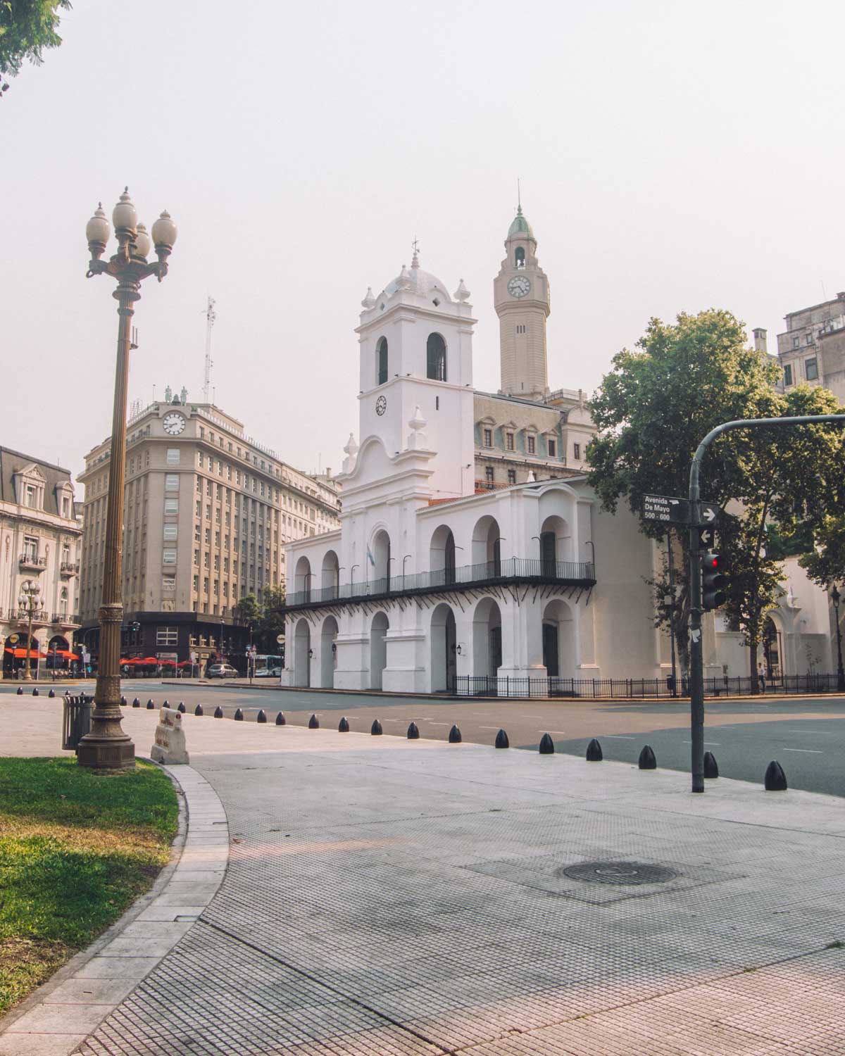 A imagem mostra o Cabildo de Buenos Aires, um edifício branco de estilo colonial, localizado na Plaza de Mayo. A arquitetura do Cabildo, com seus arcos e varanda no andar superior, se destaca no centro da cena. Ao fundo, prédios históricos e modernos coexistem, incluindo a torre do prédio com um relógio.