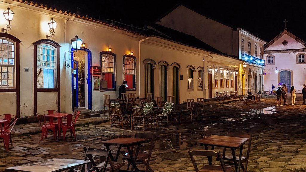 Vista de uma rua em Paraty à noite, com mesas de madeira ao ar livre, diante de edifícios coloniais iluminados. No fundo, há pessoas caminhando e uma pequena igreja ao final da rua.