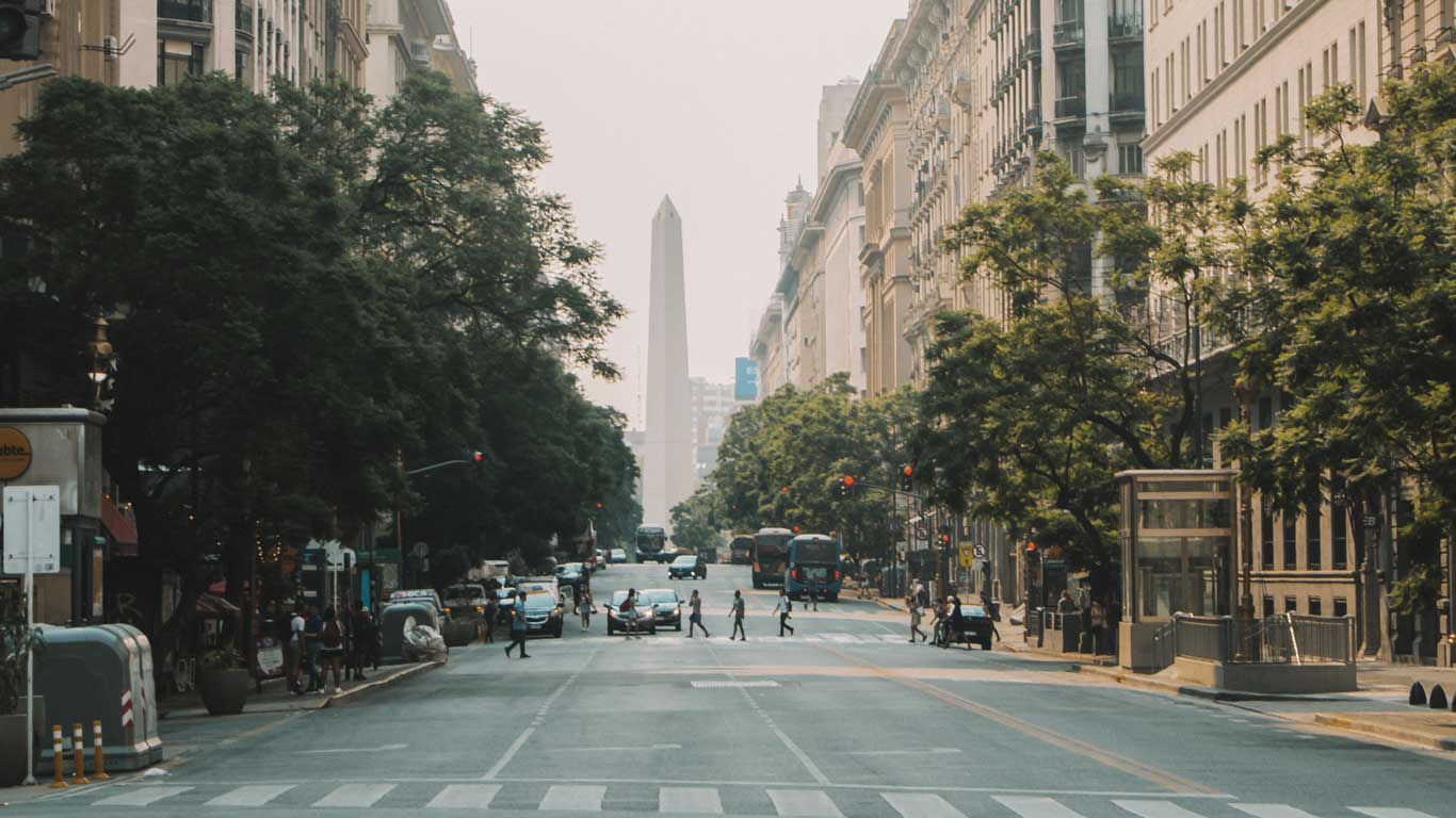 Avenida que leva ao Obelisco de Buenos Aires, um monumento icônico da Argentina. A rua é ladeada por prédios altos e árvores, e várias pessoas estão atravessando a faixa de pedestres, com tráfego leve visível. O Obelisco se destaca ao fundo, centralizado na paisagem urbana.