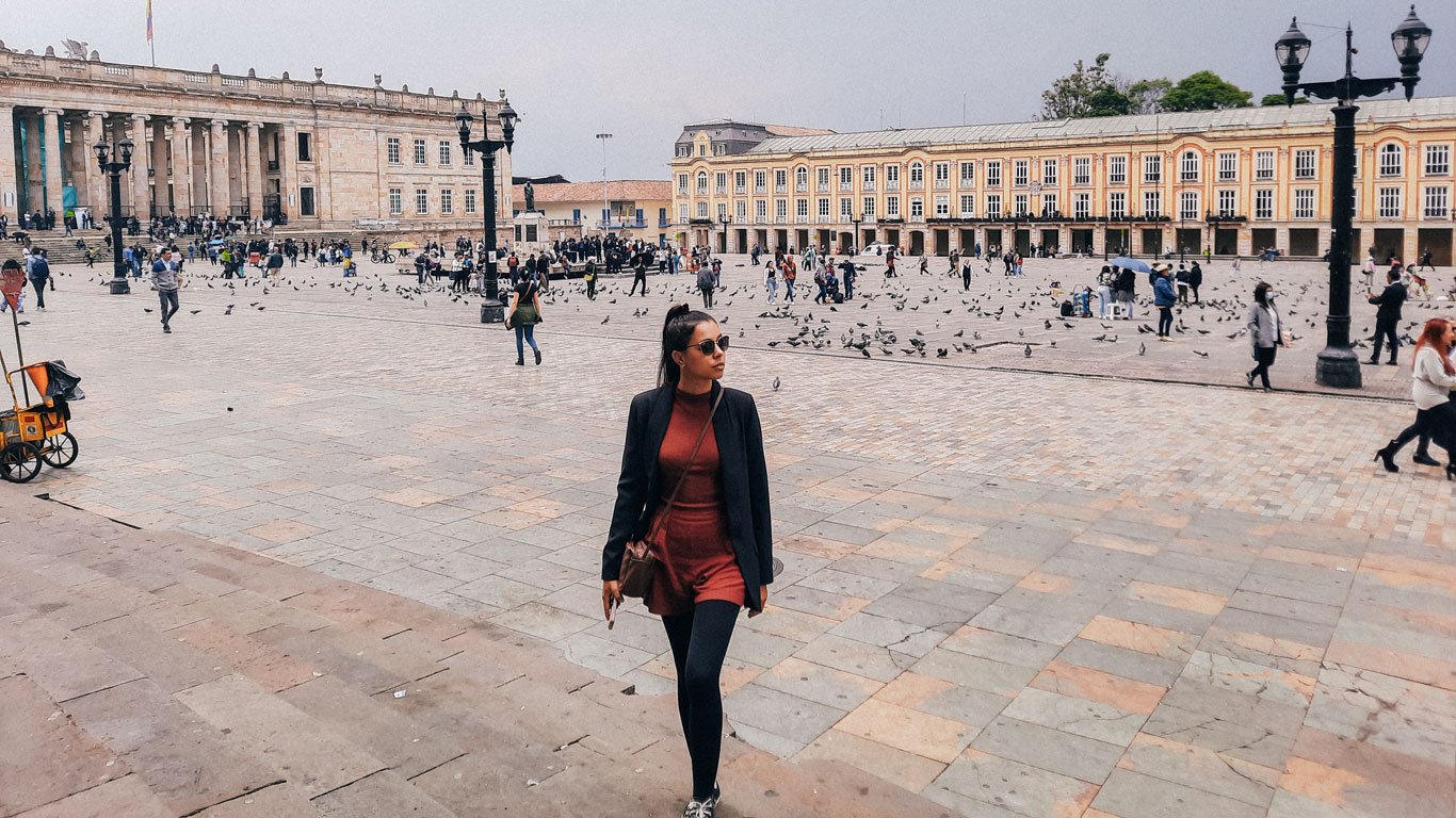 Uma mulher passeia pela ampla Praça Bolívar, rodeada por prédios históricos, incluindo o Capitólio Nacional. Pombos enchem a praça, e várias pessoas caminham e tiram fotos, aproveitando o espaço ao ar livre.