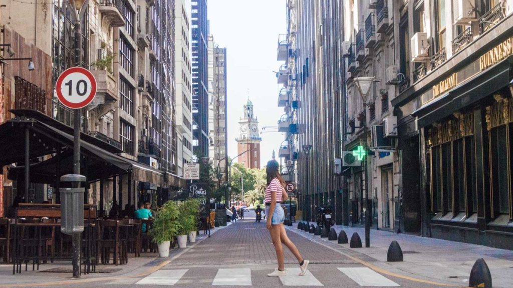 Jovem atravessando uma rua em Retiro, Buenos Aires, com o relógio da Torre dos Ingleses ao fundo. A arquitetura da região mistura edifícios históricos e modernos, e a rua, com seus pequenos restaurantes e cafés, tem um charme especial.