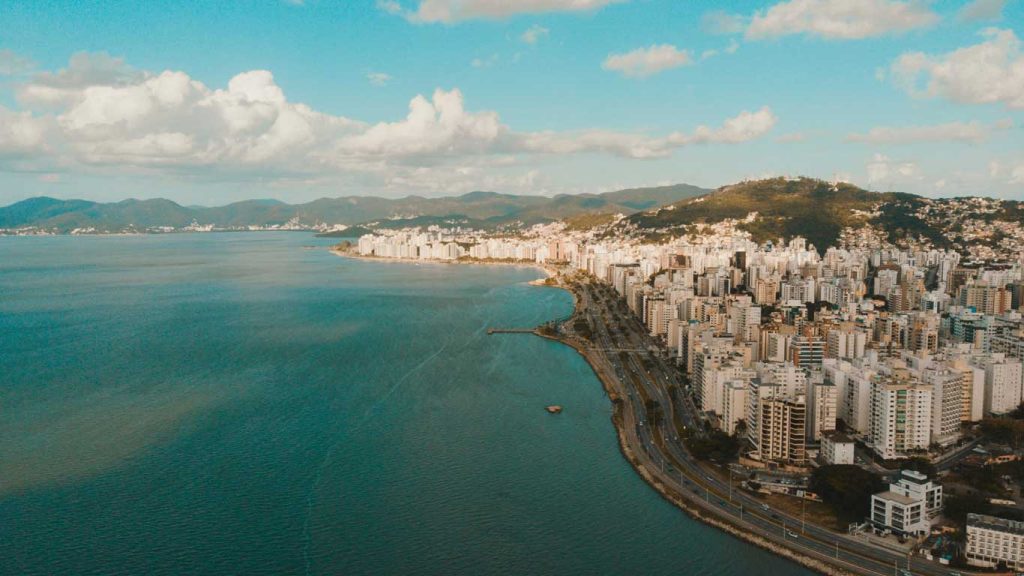 Vista aérea de Florianópolis, com prédios à beira-mar e uma estrada que acompanha a costa. Montanhas ao fundo cercam a cidade sob um céu azul com algumas nuvens.