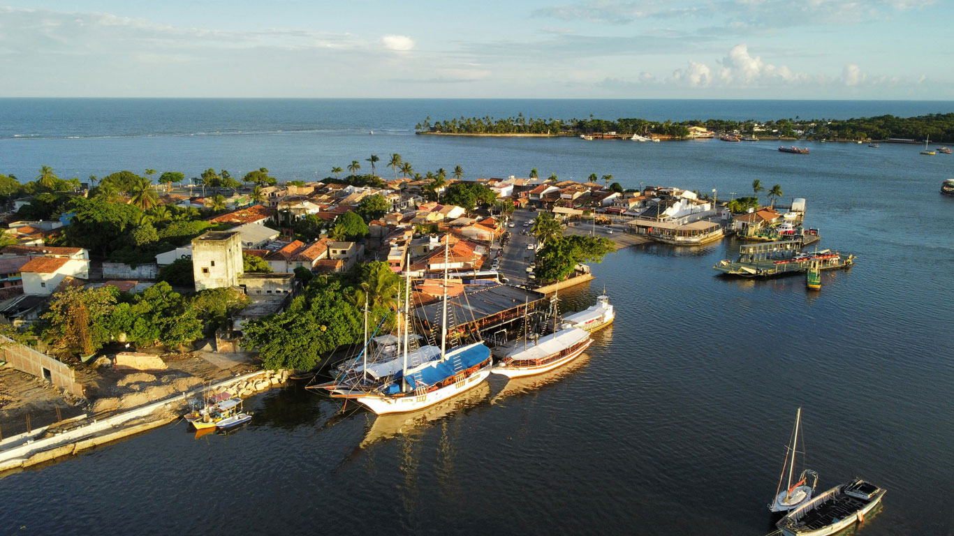 Uma vista aérea do centro de Porto Seguro, na Bahia, Brasil. Na imagem, é possível ver barcos atracados na orla e diversas construções de telhados vermelhos cercadas por vegetação tropical. Ao fundo, o mar azul e uma pequena ilha complementam a paisagem, destacando a beleza costeira e a tranquilidade do local.