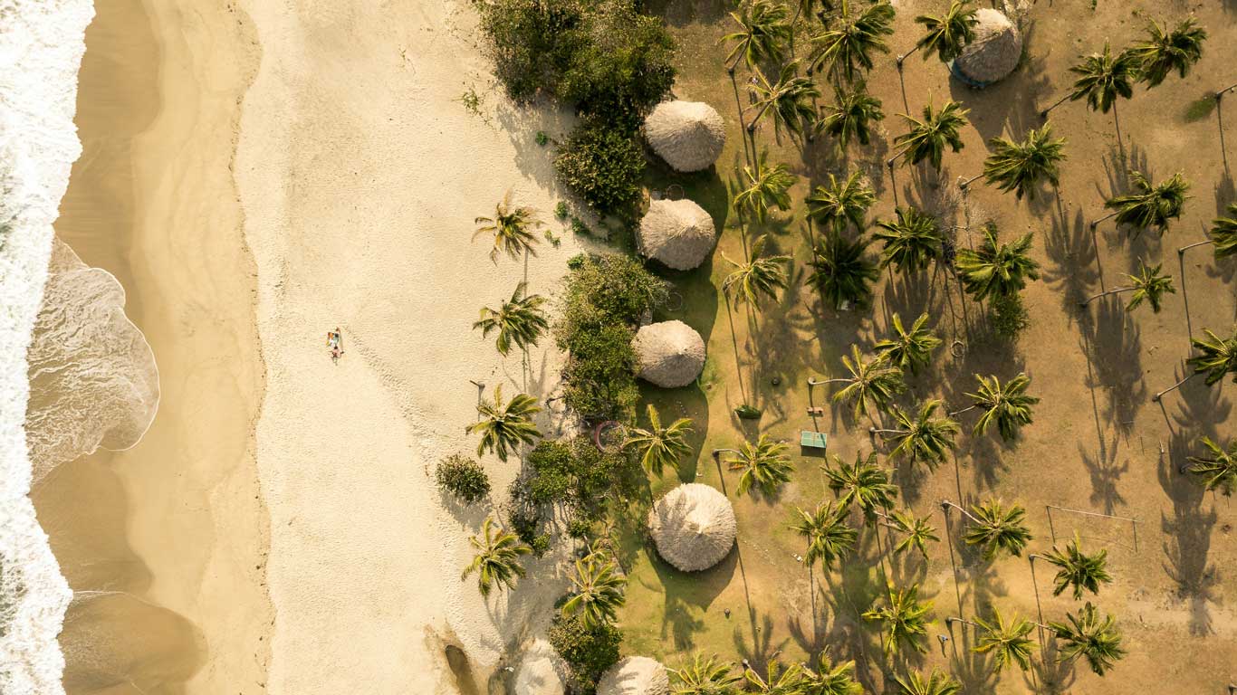 Vista aérea de uma área costeira em Tayrona, mostrando uma praia de areia dourada de um lado e coqueiros espalhados do outro. Várias cabanas com telhados de palha estão entre as palmeiras, enquanto uma pessoa está deitada sozinha na areia perto da água. Ondas suaves atingem a costa.